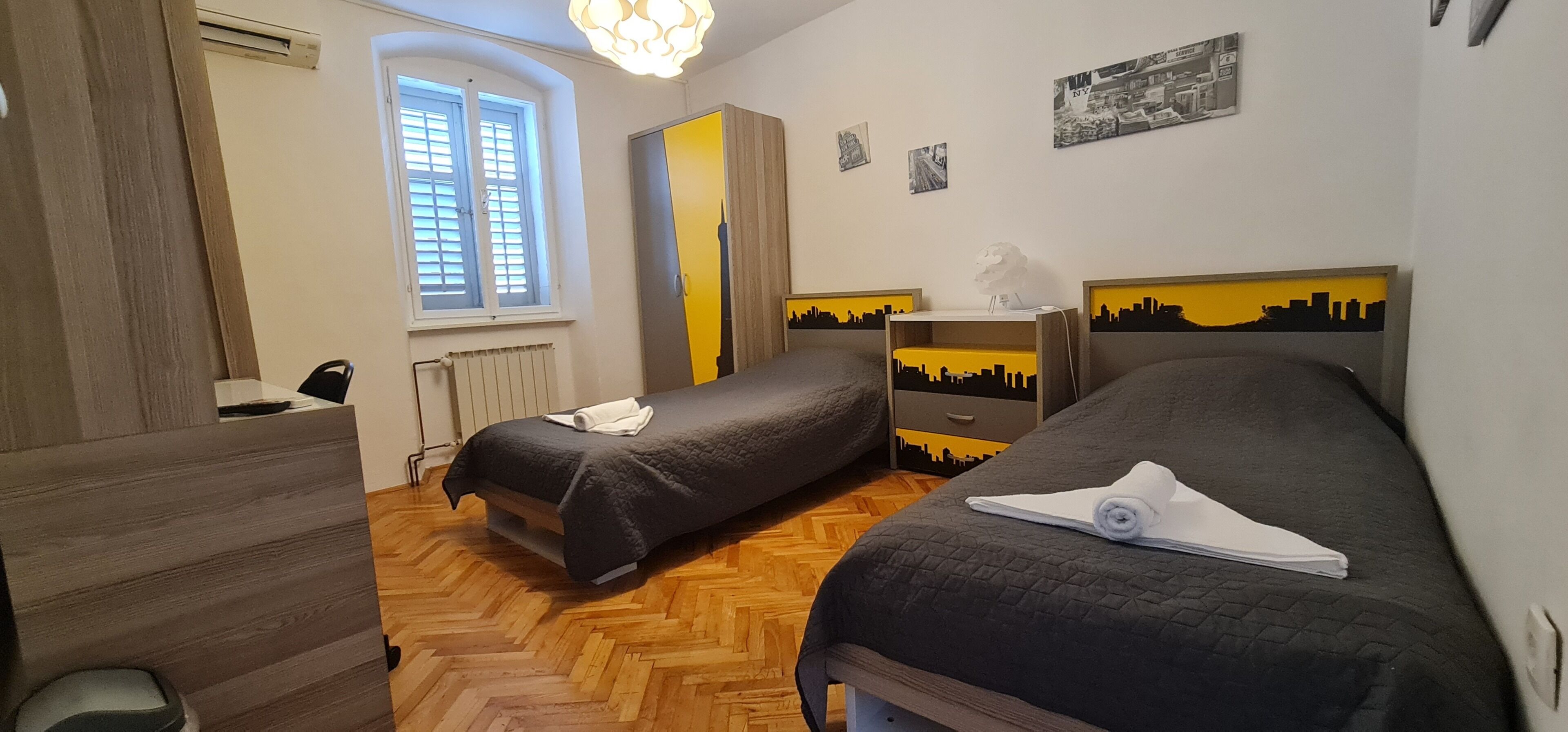 Hotelkamer met twee bedden, een houten vloer, een radiator, een raam met zonwering en een muur met stadsgezicht kunst.