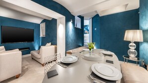 A modern living room with a flat-screen TV, a sofa, a dining table set for two, and a lamp. - Luxury 2 Bedroom Apartment in the Heart of Knightsbridge (London)
