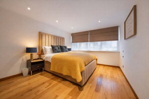 A bedroom with a bed, nightstand, lamp, and a window with blinds. - Super Deluxe Two-bedroom Apartment (London)