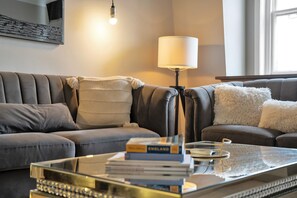 A modern living room with a leather sofa, a glass coffee table with books, a lamp, and a framed picture on the wall. - Deluxe-2bedroom Apartment Behind Harrods w AC (London)
