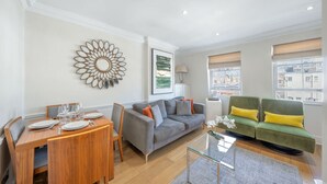 A modern living room with a dining area, a grey sofa, and a green sofa with yellow cushions. - Deluxe 2 Bedroom Apartment (London)