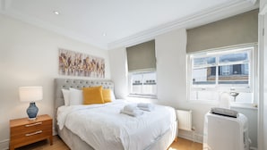 A hotel room with a bed, a nightstand, a radiator, and a window with a view of buildings. - Deluxe 2 Bedroom Apartment (London)
