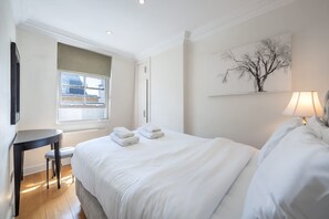 A hotel room with a bed, a window, a table, a chair, a lamp, and a painting on the wall. - Deluxe 2 Bedroom Apartment (London)