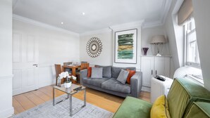 A modern living room with a grey sofa, a green armchair, and a coffee table. There is a dining area with a wooden table and chairs, and a kitchen area with a white cabinet. - Deluxe 2 Bedroom Apartment (London)