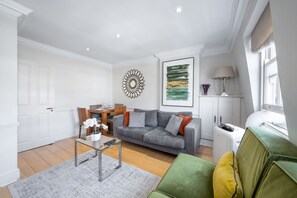 A modern living room with a grey sofa, a green armchair, and a coffee table. There is a dining area with a wooden table and chairs, and a kitchen area with a white cabinet. - Deluxe 2 Bedroom Apartment (London)