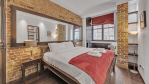 Un dormitorio con pared de ladrillo, una cama grande con ropa blanca y un plaid rojo, una mesita de noche con lámpara, una ventana con cortina roja y un espejo sobre la cama.