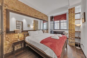 Ein Schlafzimmer mit Backsteinwand, einem großen Bett mit weißer Bettwäsche und roter Decke, einem Nachttisch mit Lampe, einem Fenster mit rotem Vorhang und einem Spiegel über dem Bett.