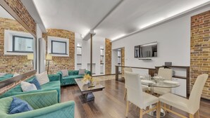Una sala de estar moderna con pared de ladrillo, sofás verdes, una mesa de comedor de vidrio y un televisor montado en la pared.