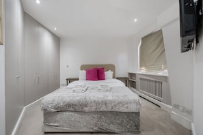 Ein Schlafzimmer mit Bett, Kopfteil, Fenster, Heizkörper und Fernseher an der Wand.