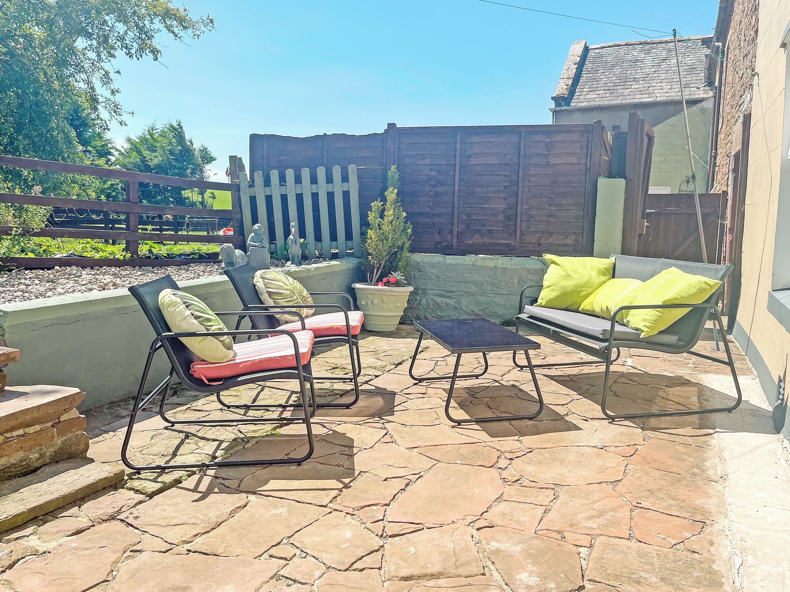 An outdoor patio with stone flooring, a wooden fence, and outdoor furniture including chairs and a sofa.