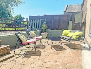 Eine Terrasse im Freien mit Steinboden, Holzlattenzaun und Gartenmöbeln, darunter Stühle und eine Couch.