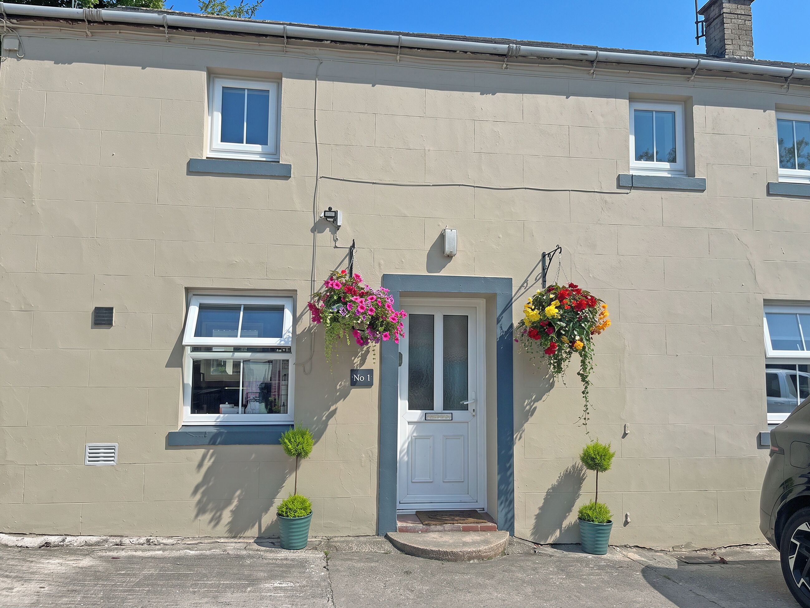 A building with a beige facade, a white door, and two windows. There are hanging flower baskets and two potted plants on the ground.