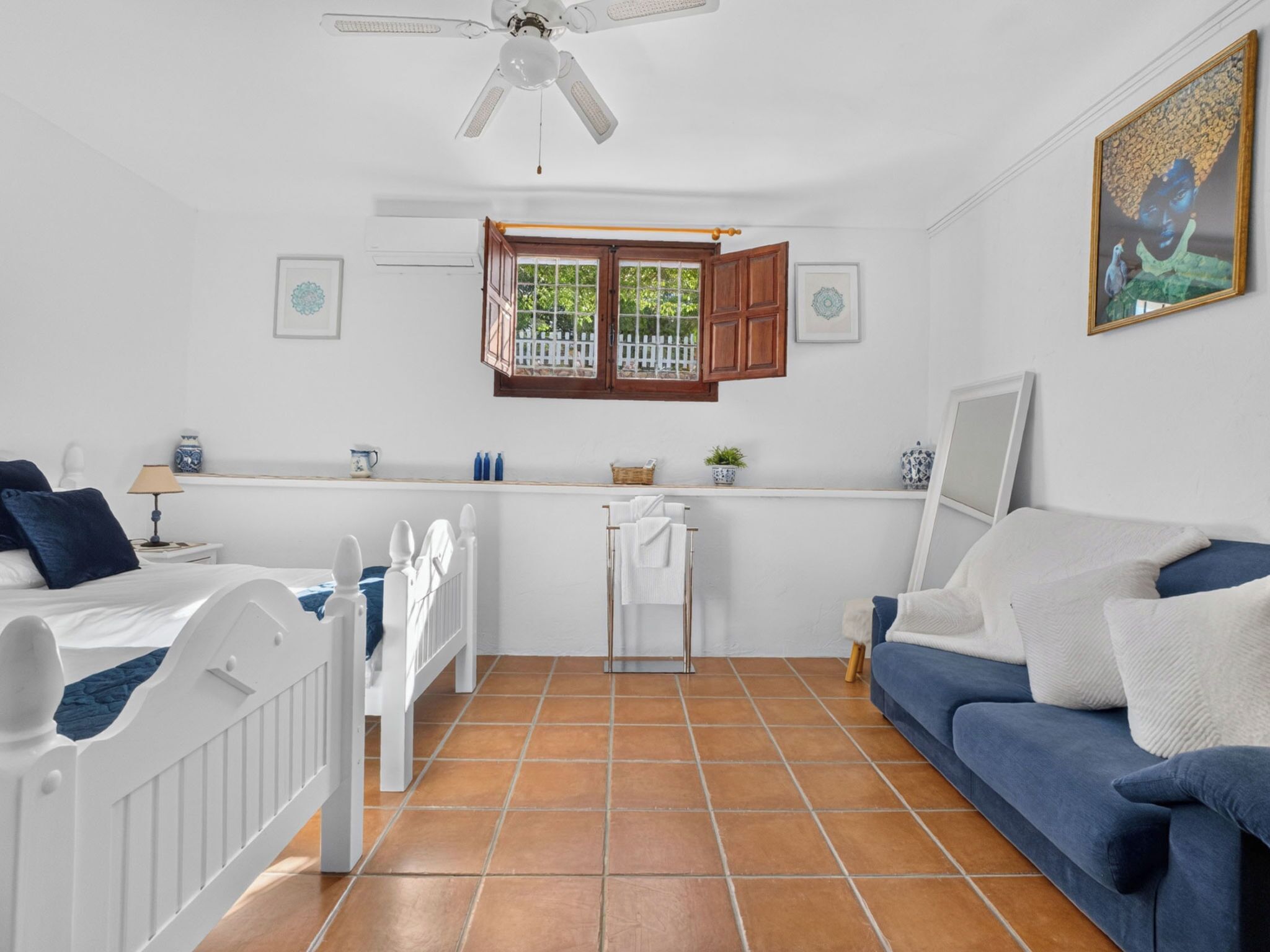 Una habitación con una cama, un sofá azul, un radiador blanco, una ventana con persianas de madera y un ventilador de techo.