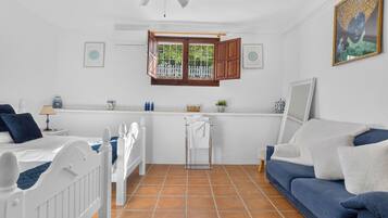 Una habitación con una cama, un sofá azul, un radiador blanco, una ventana con persianas de madera y un ventilador de techo.