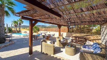 Een terras buiten met rieten meubels, een zwembad en een pergola.