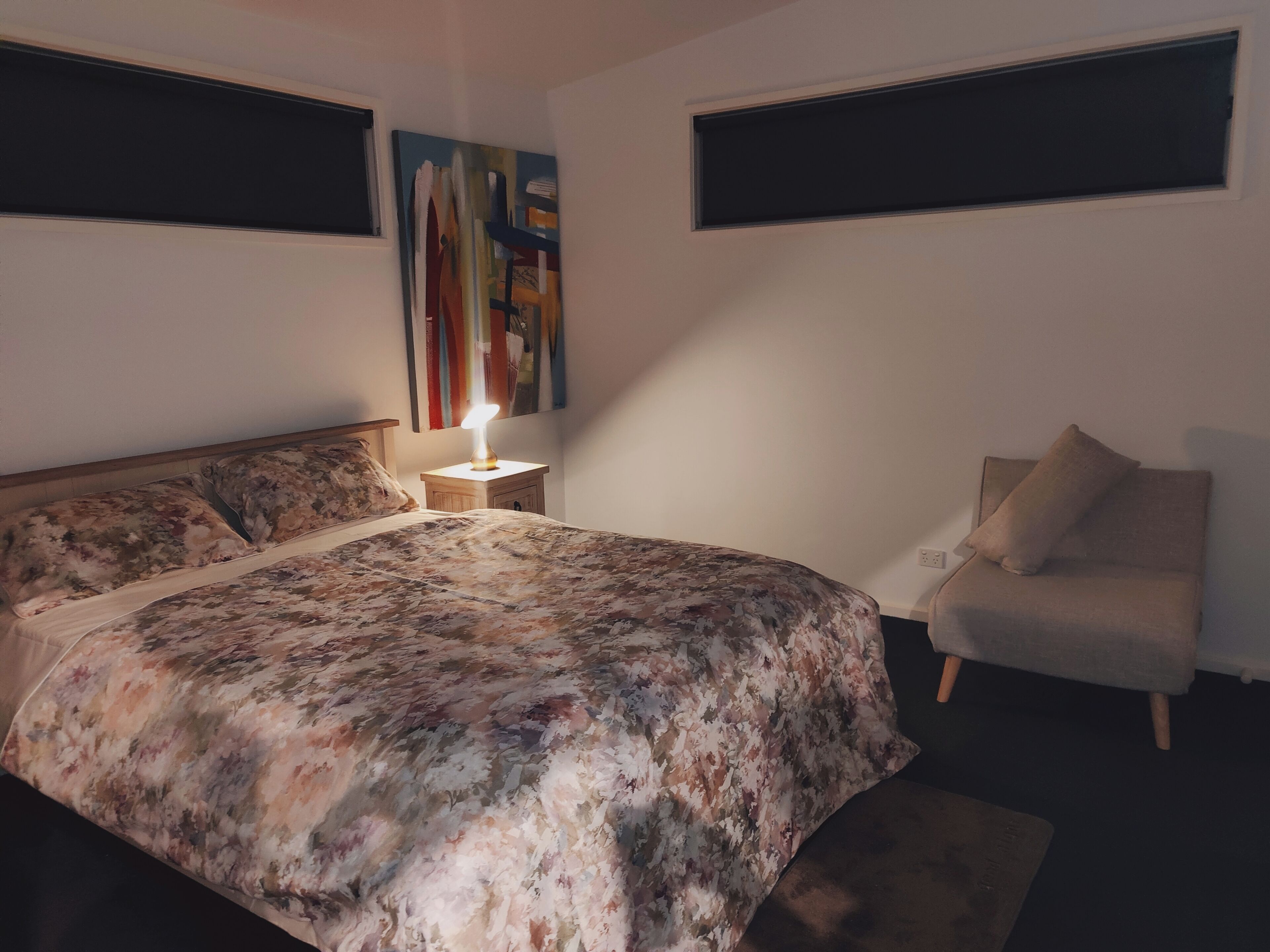 A bedroom with a bed, nightstand, lamp, chair, and artwork on the wall.