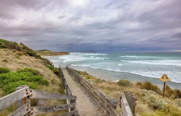 A wooden boardwalk leading to a beach with ocean views, a sign indicating a 50-meter distance, and a cloudy sky.