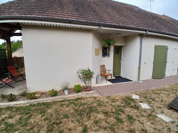 Um edifício com porta verde, um pequeno alpendre com uma cadeira e uma área coberta com dois assentos.