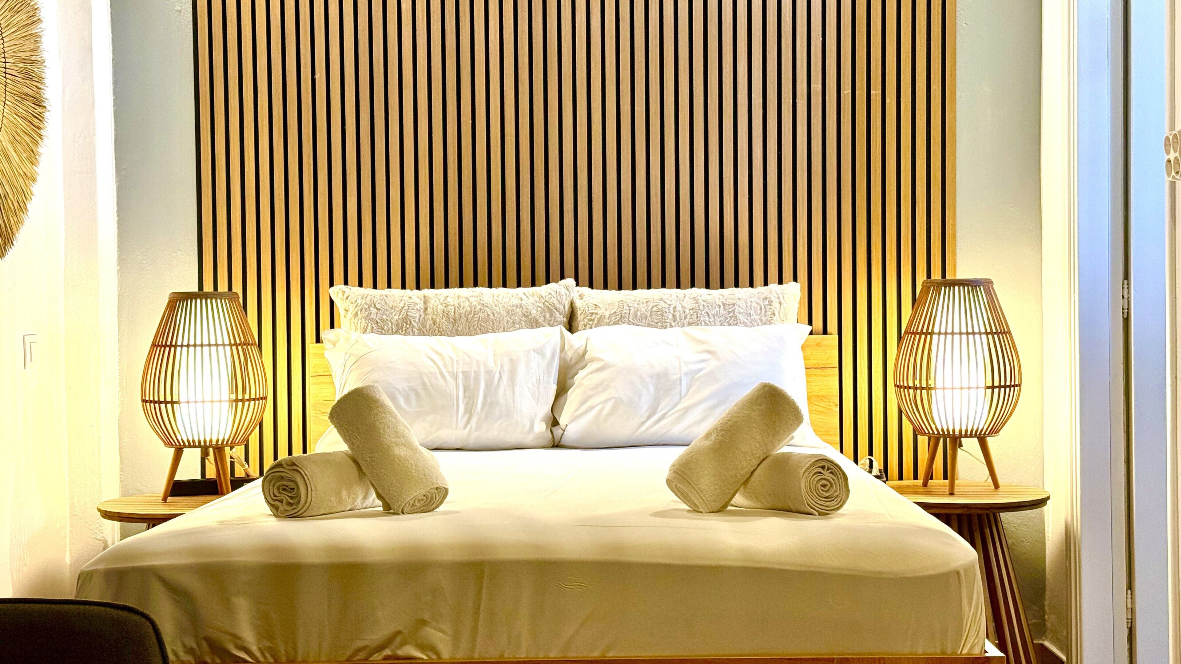 A neatly made bed with pillows and rolled towels, two bedside lamps, and a wooden slat headboard.