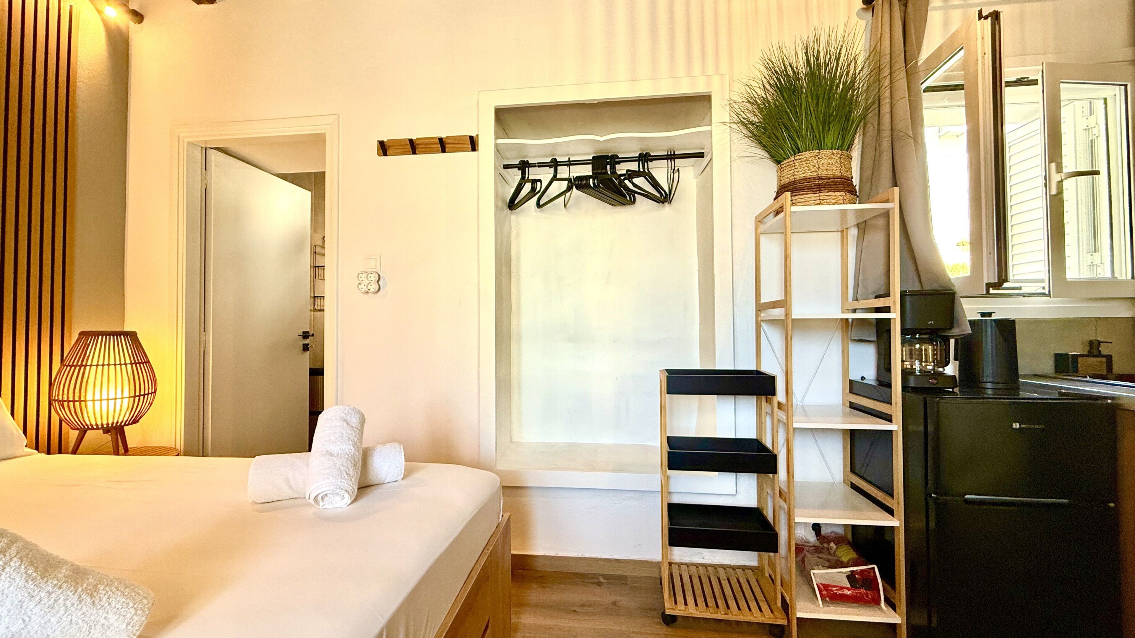 A hotel room with a bed, a wooden shelf with a plant, a coat rack, and a minibar.