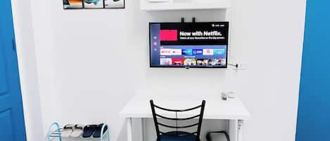 Ein Zimmer mit weißem Schreibtisch, blauem Stuhl, Wand-Fernseher, Ablage mit Mikrowelle und Paar Hausschuhen auf einem Ablagegestell.