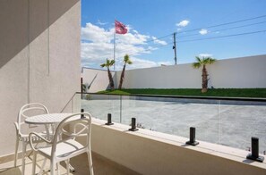 A balcony with a white chair and a table, overlooking a grassy area with palm trees and a flag.