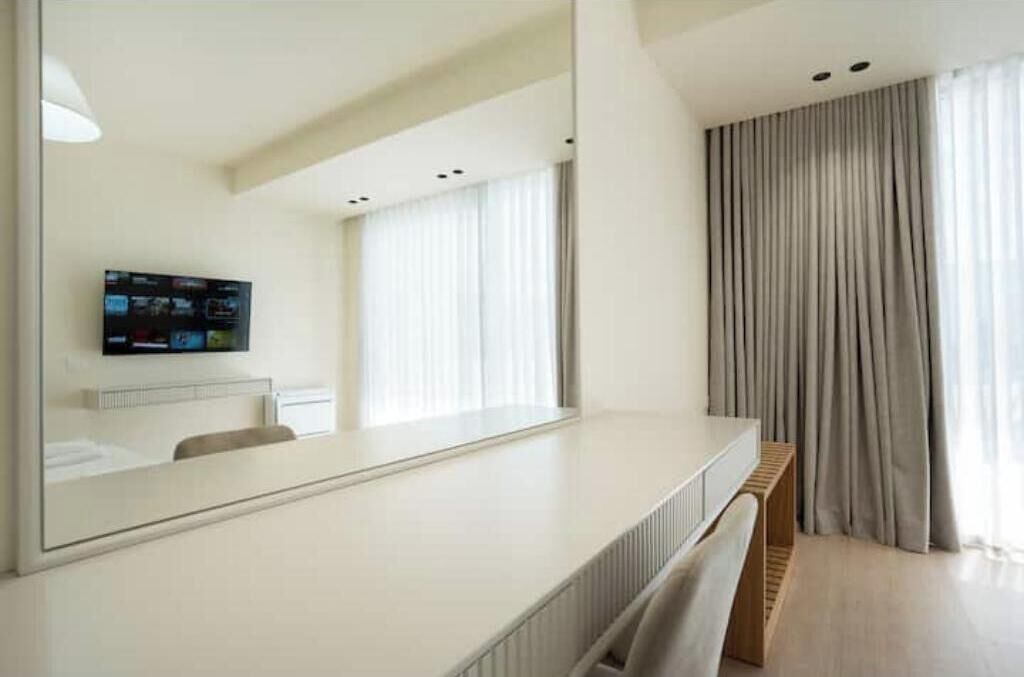 A modern hotel room with a large mirror, a flat-screen TV mounted on the wall, a wooden bench, and a large window with curtains.