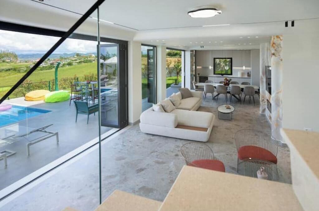 A modern living room with a large glass wall, a white sofa, and a dining area with chairs.