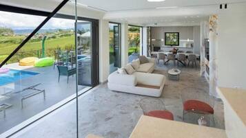 A modern living room with a large glass wall, a white sofa, and a dining area with chairs.
