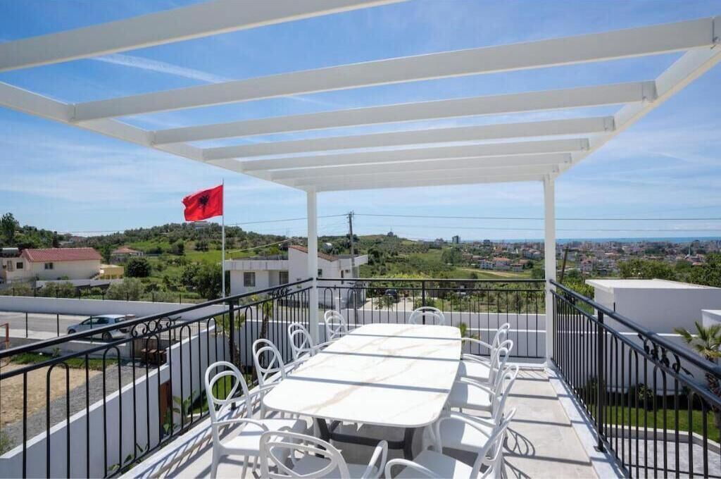 A balcony with a white pergola, a table with chairs, and a flag.