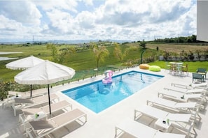 A swimming pool with lounge chairs and umbrellas, surrounded by a grassy area and a view of the countryside.
