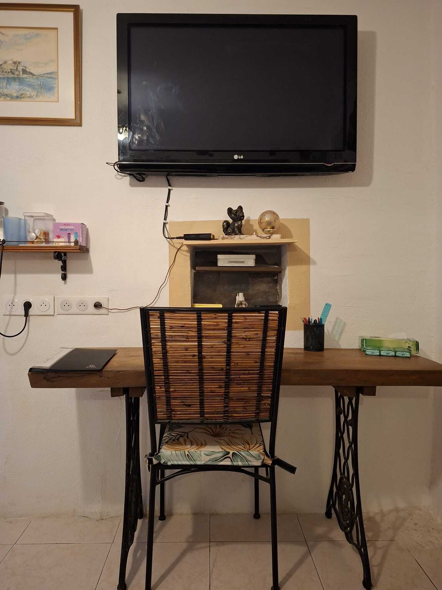 Quarto com televisão de tela plana na parede, uma escrivaninha de madeira com cadeira e uma prateleira com diversos objetos.
