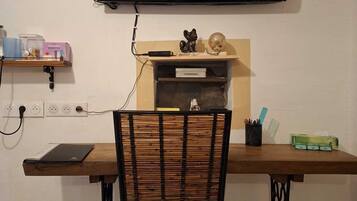 Quarto com televisão de tela plana na parede, uma escrivaninha de madeira com cadeira e uma prateleira com diversos objetos.
