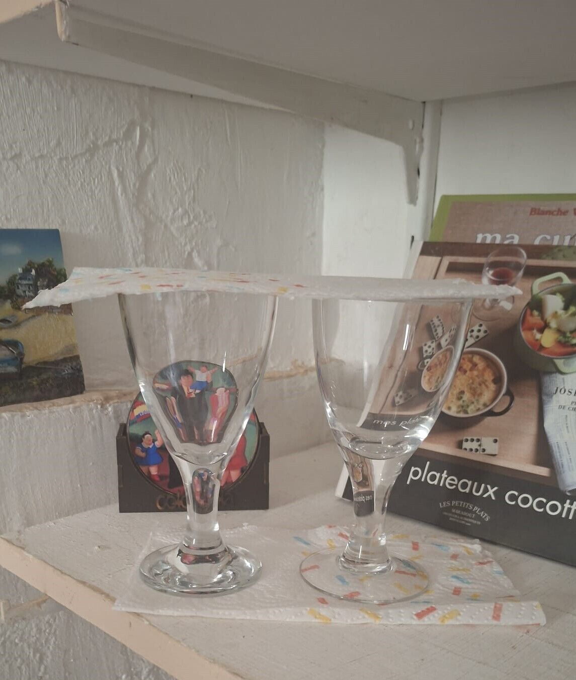 Two clear glass wine glasses on a table with a white tablecloth, a box of 'plateaux cocotte' in the background, and a small decorative item with a picture.