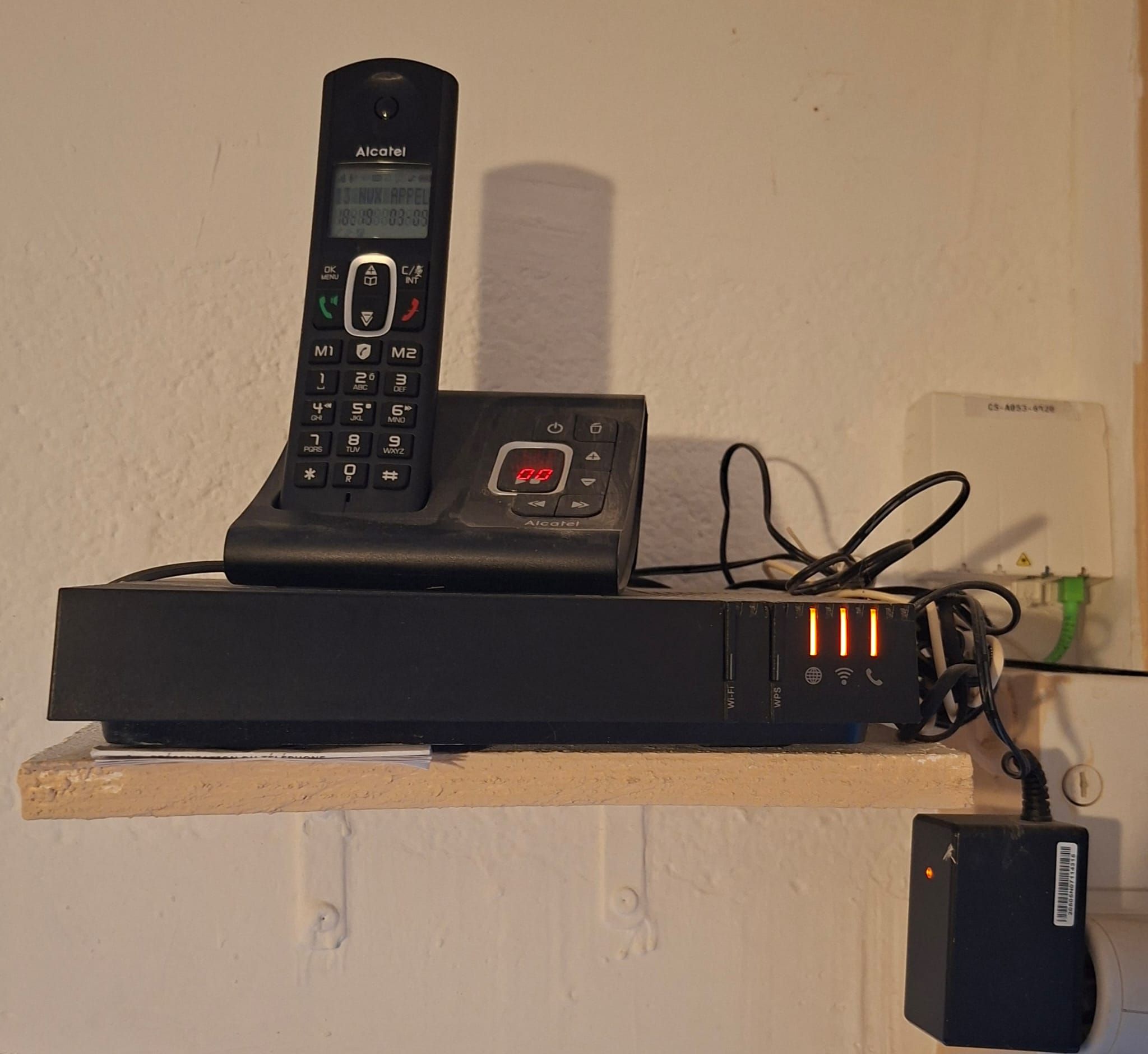 A black Alcatel phone on a wooden shelf with a power strip and cables.