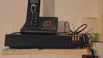 A black Alcatel phone on a wooden shelf with a power strip and cables.