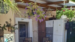 Terrassa exterior amb taula i cadires de fusta, envoltada de plantes penjants i un frigorífic.