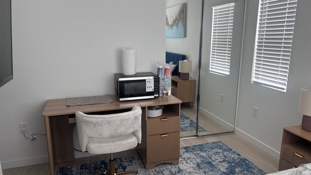 A bedroom with a wooden desk, a chair, a bed, a mirror, and a window with blinds. - Peaceful brand new home in a gated community in Azul (Pico Rivera)
