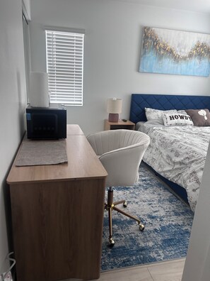 Ein Schlafzimmer mit einem Bett, einem Schreibtisch, einem Stuhl, einem Nachttisch, einer Lampe und einem Bild an der Wand.