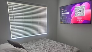 A bedroom with a bed, a window with blinds, and a TV displaying a QR code. - Peaceful brand new home in a gated community in Azul (Pico Rivera)