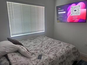 Ein Schlafzimmer mit einem Bett, einem Fenster mit Jalousien und einem Fernseher, der einen QR-Code zeigt.