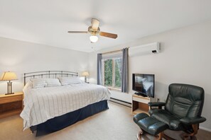 Camera da letto con un letto, un ventilatore a soffitto, una TV, una sedia, una finestra con tende e un comodino con una lampada.