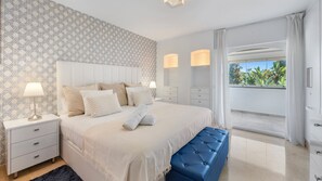 Une chambre à coucher avec un lit, des tables de chevet, un banc et un balcon agrémenté de rideaux.