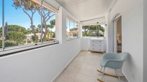 Un balcon donnant sur des arbres et des immeubles, une commode blanche et un fauteuil à bascule.