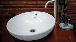 Un lavamanos blanco con un grifo moderno, un estante pequeño con tres botellas, y una pared de madera al fondo.