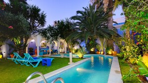 Piscina con sillones de descanso, palmeras y un jardín bien iluminado.