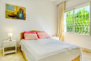 Une chambre à coucher avec un lit, une table de chevet, une lampe, une fenêtre donnant sur un paysage verdoyant et un tableau accroché au mur.