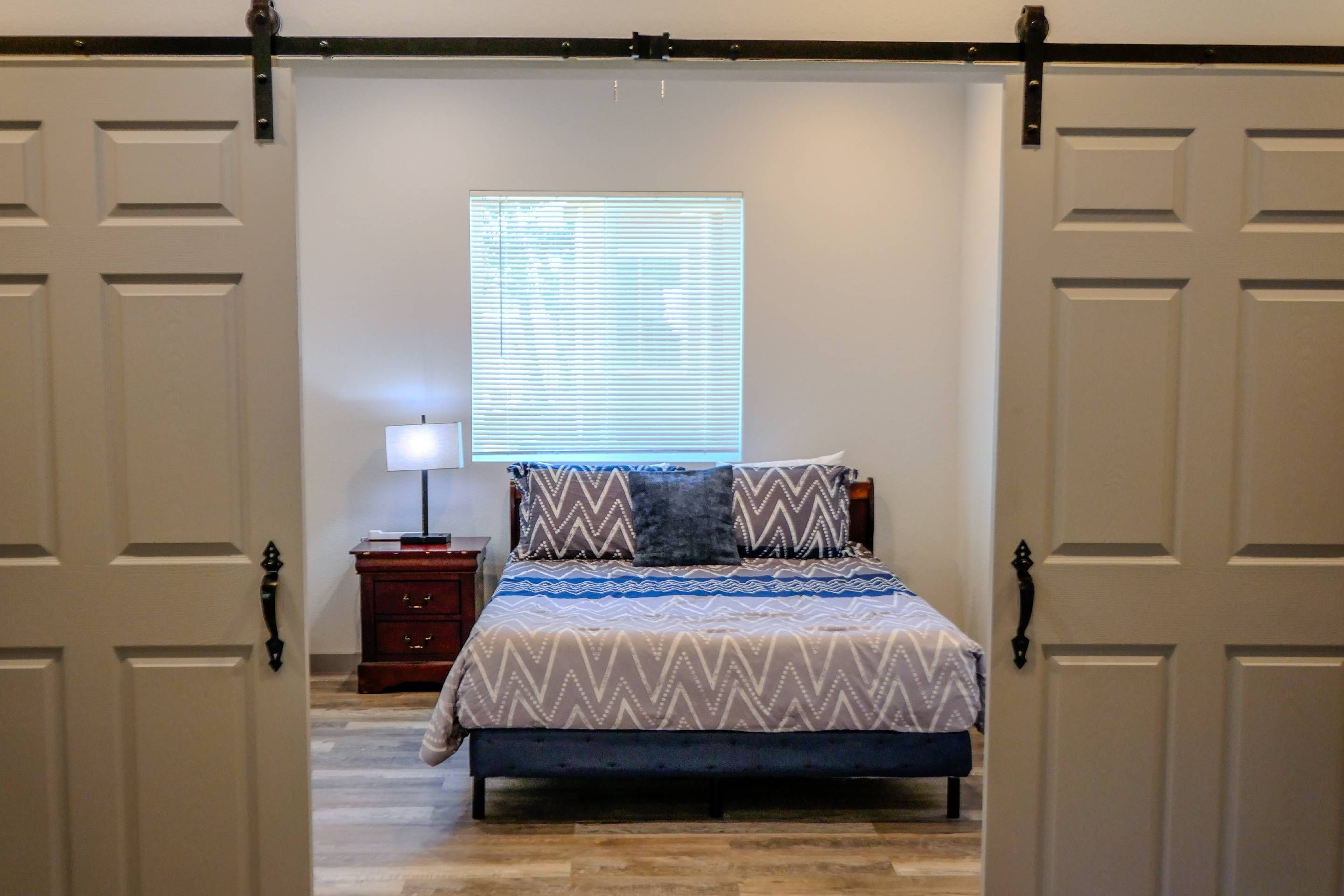 Un dormitorio con cama, mesita de noche, lámpara, ventana con persianas y puerta con sistema corredizo.