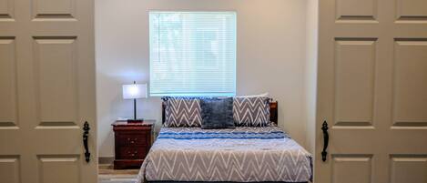 Un dormitorio con cama, mesita de noche, lámpara, ventana con persianas y puerta con sistema corredizo.