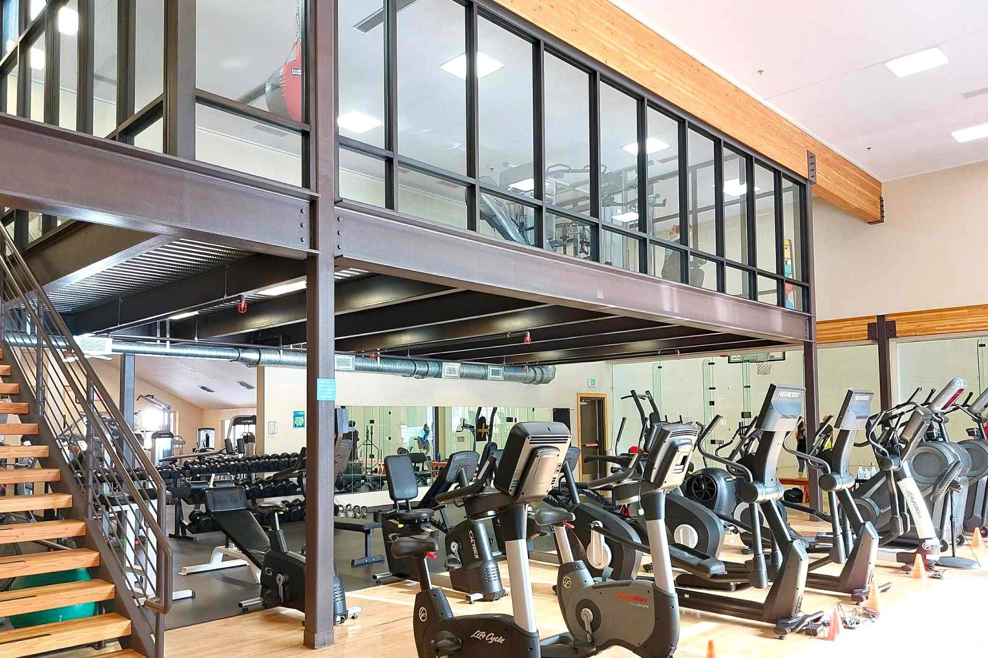 A gym with multiple exercise machines, a staircase, and a mezzanine level with large windows.